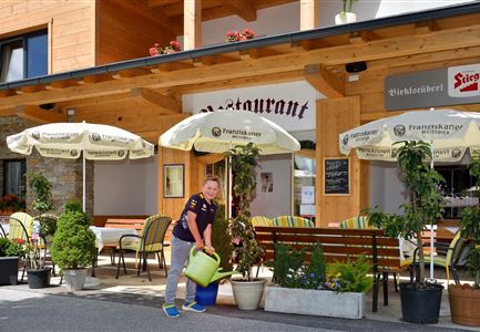 Ein Restaurant mit einer einladenden Terrasse und Sonnenschirmen. Jemand gießt die Pflanzen vor dem Eingang.
