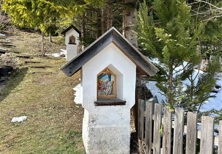 A small chapel with a painted image stands by a path in nature. In the background, trees and some snow can be seen.