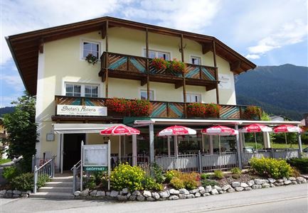 Ein charmantes Gasthaus mit mehreren Balkonen, umgeben von bunten Blumen. Vor dem Gebäude befinden sich Sonnenschirme und eine gemütliche Terrasse.