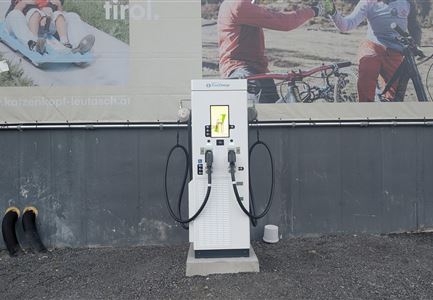 Eine E-Ladesäule steht vor einer Wand mit Werbeanzeigen. Daneben befinden sich einige Gummistiefel auf dem Boden.