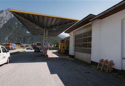 A gas station with a large, shade-providing roof. In the background, mountains and a building are visible.