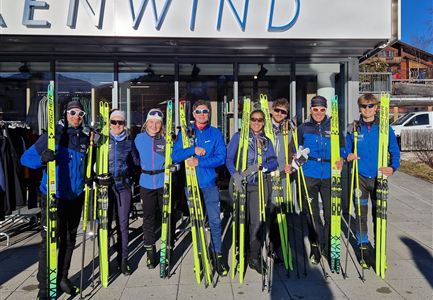 Eine Gruppe von acht Skifahrern in blauen Outfits steht vor einem Geschäft. Jeder hält ein Paar Skier in der Hand, und die Sonne scheint.