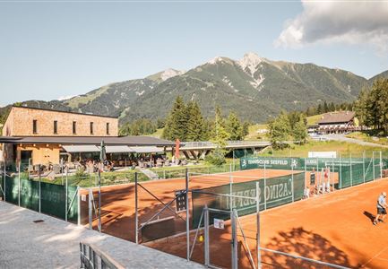 Eine Tennisanlage mit roten Sandplätzen und einem modernen Gebäude. Im Hintergrund sind beeindruckende Berge und eine grüne Landschaft zu sehen.
