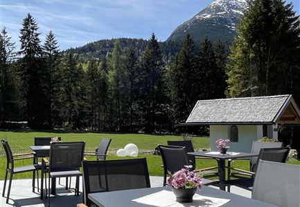 Eine Terrasse mit Tischen und Stühlen umgeben von einem grünen Garten. Im Hintergrund sind Bäume und ein Berg mit schneebedecktem Gipfel zu sehen.
