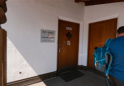 Eine Tür zu einem WC mit einem Informationsschild daneben. Im Hintergrund ist eine Person mit einem Rucksack zu sehen.
