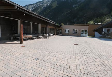 A paved courtyard with several buildings in the background. The surroundings are mountainous and green.