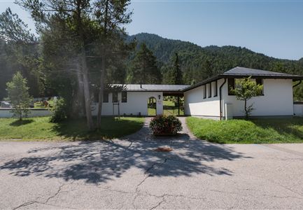 A modern house with a flat roof and generous driveway. Behind the house, trees and hills are visible.
