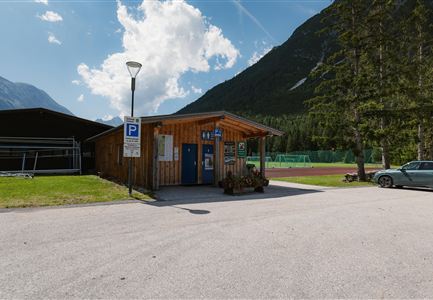 Ein Holzgebäude mit Parkplatz und Sportplätzen im Hintergrund. Die Umgebung ist grün und von Bergen umgeben.