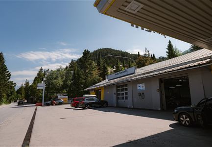 Eine Werkstatt umgeben von Bäumen und Bergen. Einige Autos stehen auf dem Parkplatz.