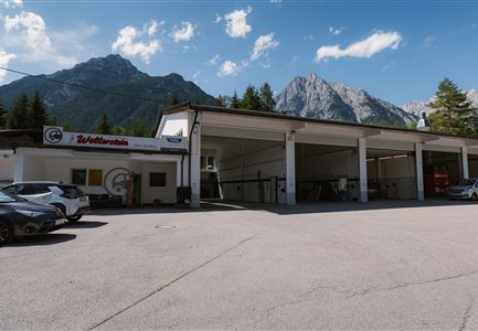 Eine Werkstatt mit modernen Gebäuden und mehreren Autos vor der Tür. Im Hintergrund sind beeindruckende Berge und ein blauer Himmel zu sehen.