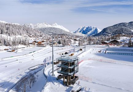 A picturesque winter landscape with snow-covered mountains and a ski area. In the foreground stands a modern building, surrounded by snowy slopes and skiers.