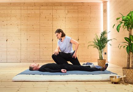 Eine entspannende Yoga-Sitzung in einem lichtdurchfluteten Raum. Eine Person zeigt eine Übung auf einer Matte, während eine andere Person entspannt liegt.