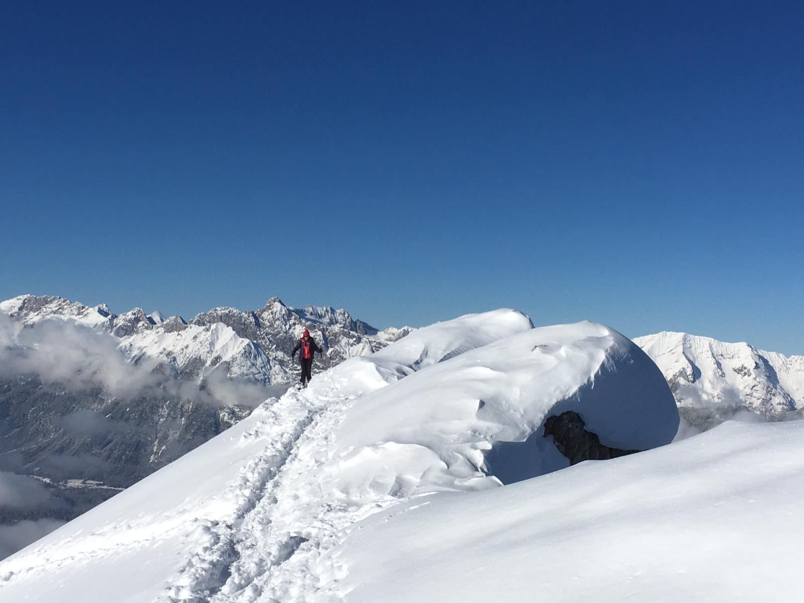 Eine verschneite Berglandschaft unter einem klaren blauen Himmel. Im Vordergrund sieht man Fußspuren im Schnee und einen Wanderer auf dem Gipfel.
