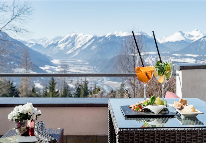 Een terras met uitzicht op majestueuze bergen. Op de tafel staan twee kleurrijke cocktails en een kleine snack om van te genieten.