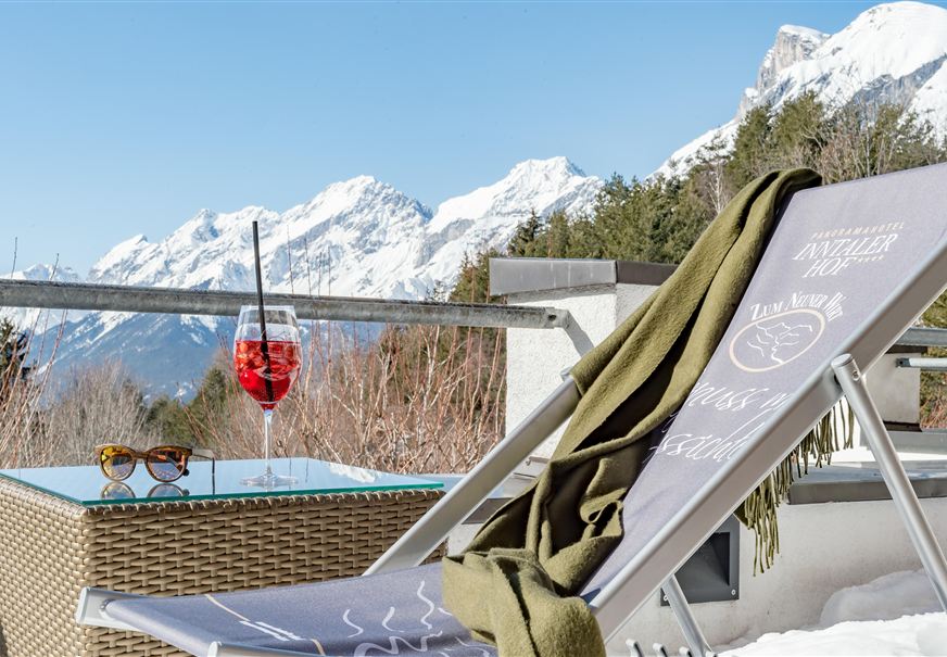 Een ligstoel met een drankje staat op een terras met uitzicht op besneeuwde bergen. Naast de ligstoel ligt een warme deken en een zonnebril.