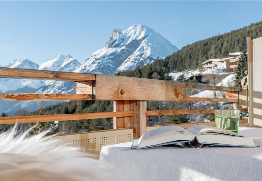 Eine gemütliche Terrasse mit Blick auf schneebedeckte Berge. Ein geöffnetes Buch und ein Glas Wasser stehen auf dem Tisch.