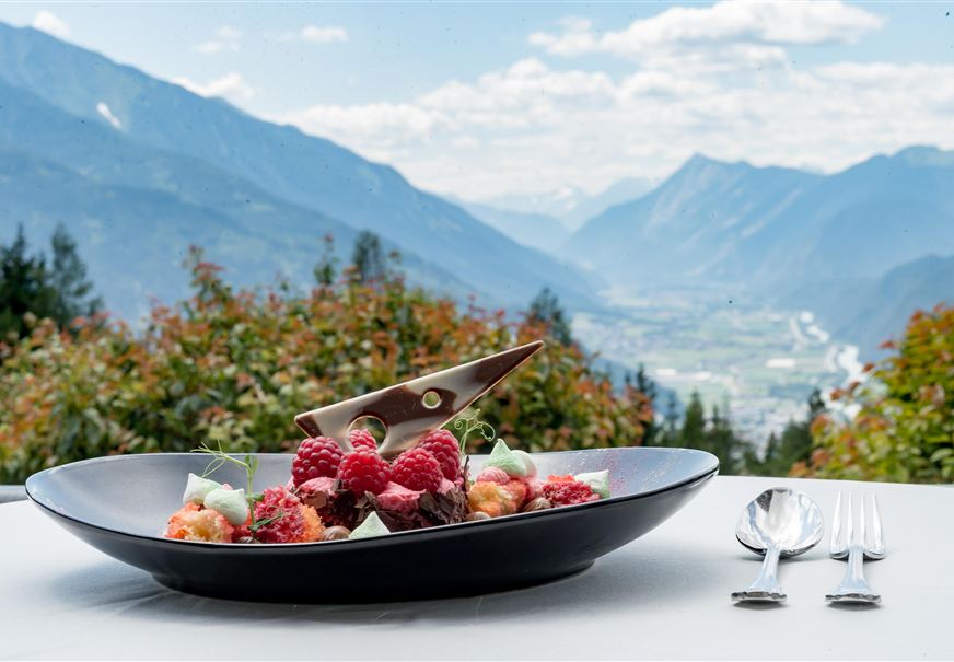 Een elegant dessert op een zwart bord, omringd door verse frambozen en decoratieve elementen. Op de achtergrond strekt zich een schilderachtig berglandschap uit met een heldere lucht.