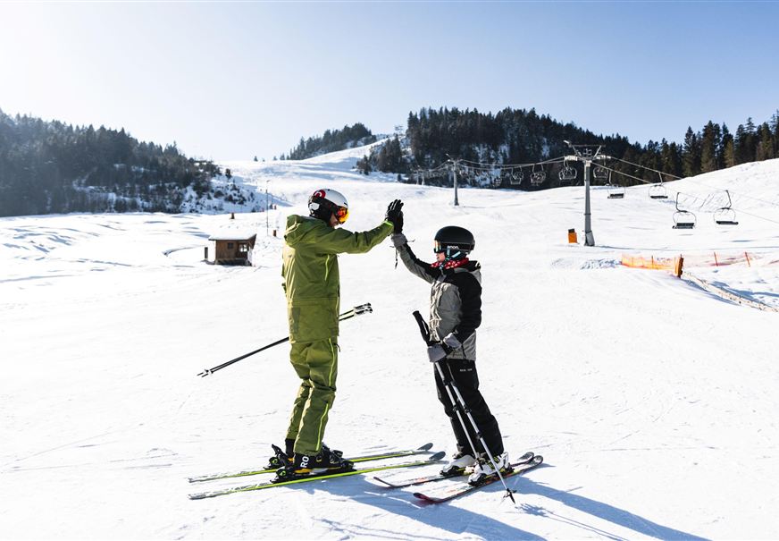 Zwei Skifahrer feiern einen erfolgreichen Moment auf der Piste. Im Hintergrund sind schneebedeckte Berge und ein klarer blauer Himmel zu sehen.