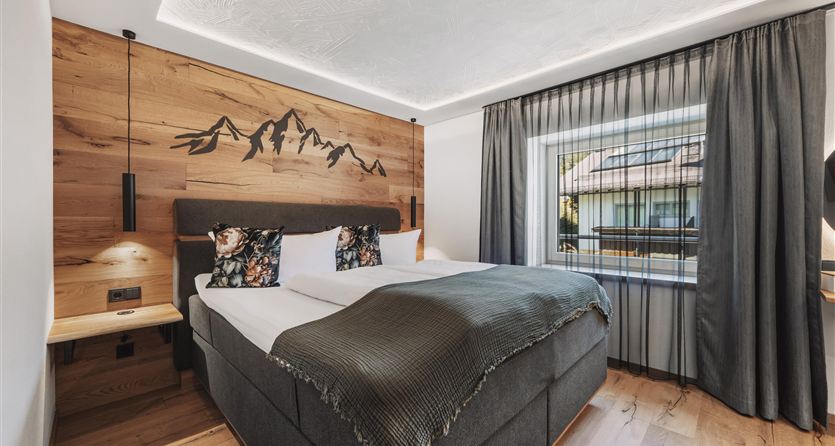 A modern bedroom with a comfortable double bed and wooden details. Large windows let in plenty of light and offer a view of the surroundings.