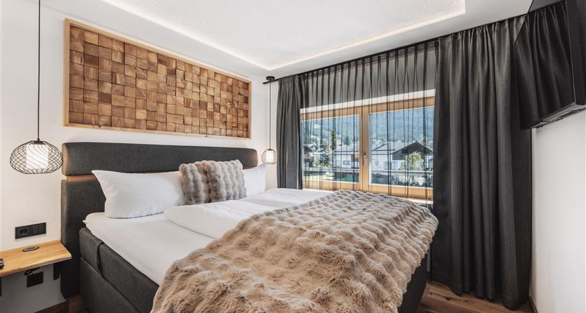 A modern bedroom with a large bed and a cozy blanket. Large windows offer a view of the surroundings and let in plenty of light.