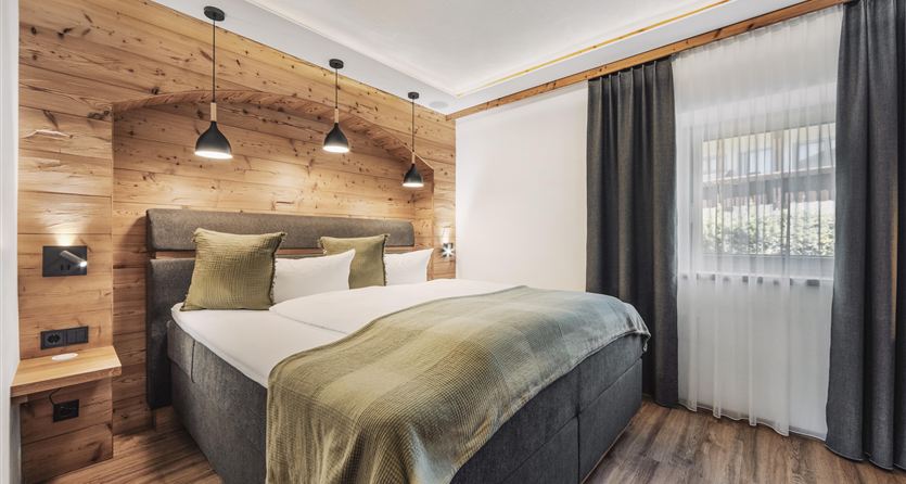 A modern bedroom with wooden panels and a large double bed. The soft lighting and the curtains create a cozy atmosphere.