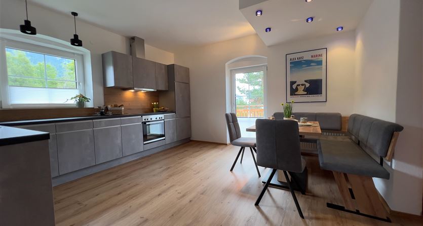 A modern kitchen with gray cabinets and a dining area. The room has large windows that let in plenty of light.