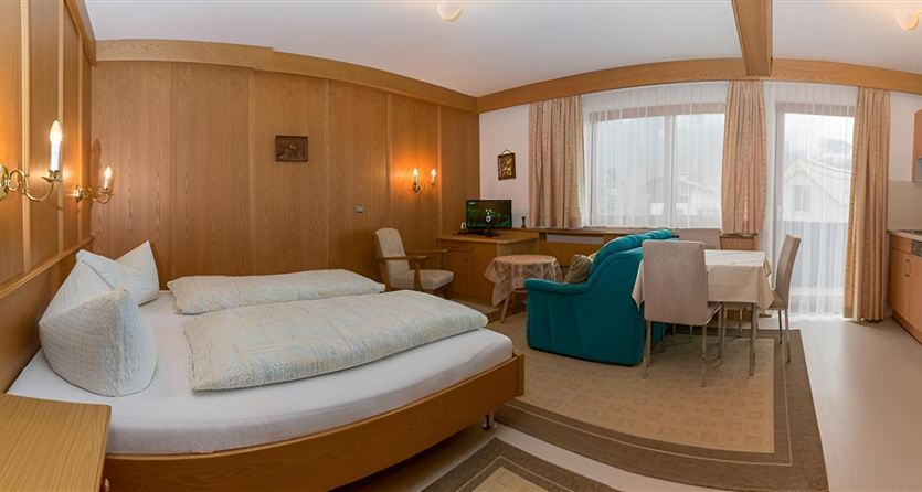 A cozy room with a double bed and wooden paneling. There is a seating area, a table, and a kitchenette.