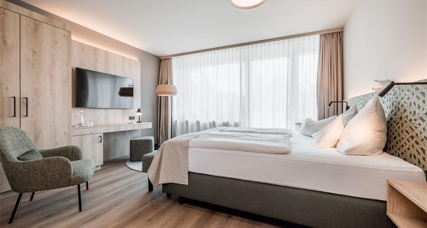 A modern hotel room with a large bed and soft colors. There is a desk, a television, and plenty of natural light through the windows.