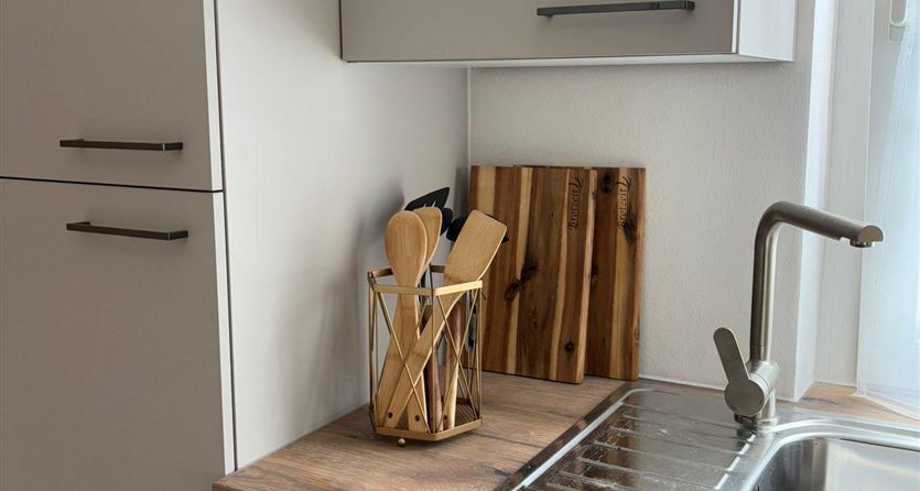 A modern kitchenette with a stainless steel sink and wooden countertop. Next to the sink, there is a container with cooking utensils and a wooden cutting board on the work surface.