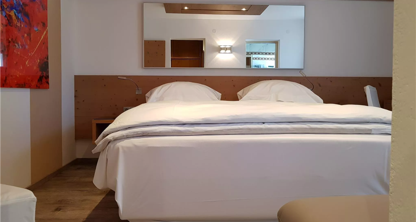 Een gezellige slaapkamer met een groot bed en lichte dekens. Aan de muur hangt een kleurrijk schilderij en een spiegel weerkaatst het licht.