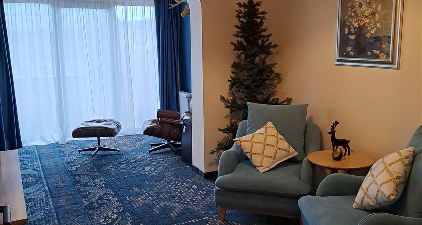 A cozy living space with two blue armchairs and a Christmas tree in the corner. Curtains reveal a light-filled room and a rug with an appealing pattern.