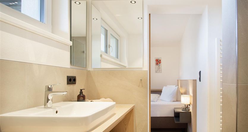 Een modern badkamer met een wastafel en grote spiegels. Op de achtergrond is een lichte slaapruimte zichtbaar.