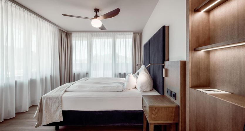 A modern bedroom with a large bed and a wooden shelf.  
Large windows let in plenty of daylight.
