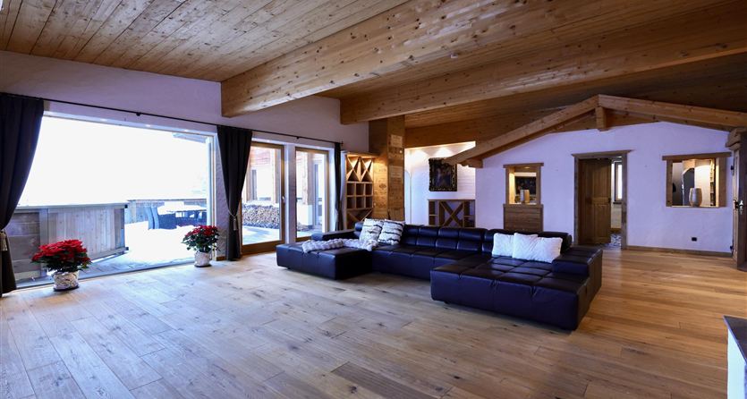 Ein modernes Wohnzimmer mit Holzboden und einer großen Fensterfront. Die Sitzecke ist mit einem schwarzen Sofa und hellen Kissen ausgestattet.