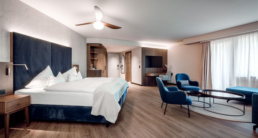 A modern hotel room with a large bed and two blue chairs.  
Large windows provide plenty of daylight and a welcoming atmosphere.
