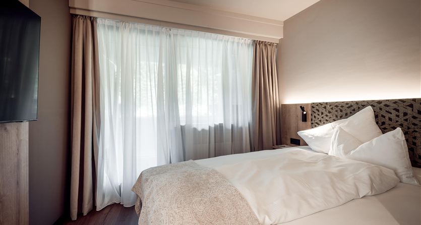 A modern bedroom with a large window and daylight. It is elegantly furnished with white bedding and a TV on the wall.