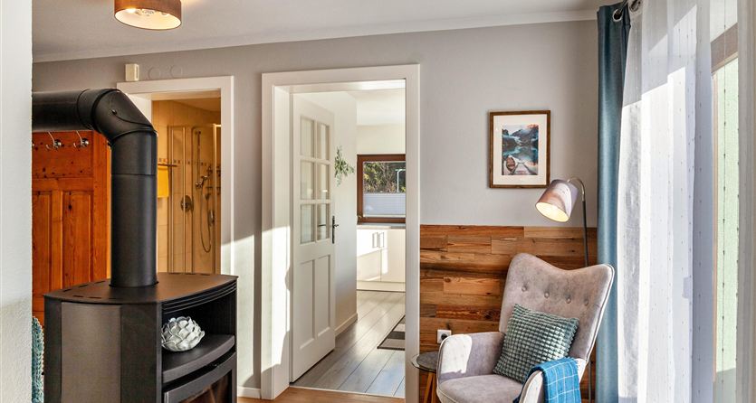 Un accogliente soggiorno con una poltrona e una stufa a legna. L'area aperta conduce a una cucina luminosa con una grande finestra.