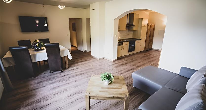 Een moderne woonkamer met een grijze bank en een houten tafel. Op de achtergrond ziet men een eettafel en een open keuken.