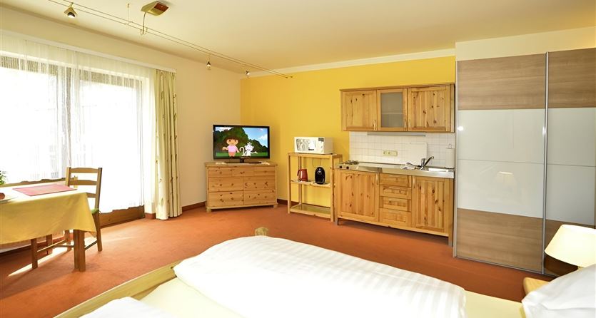Ein gemütliches Zimmer mit einem großen Bett und einer modernen Küchenecke. Es gibt einen Fernseher und viel Tageslicht durch die Fenster.