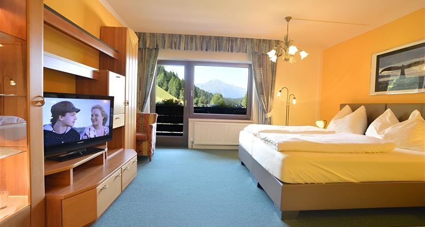 Ein helles Hotelzimmer mit einem großen Fenster und Blick auf die Berge. Es gibt ein Doppelbett, einen Fernseher und einen gemütlichen Sessel.
