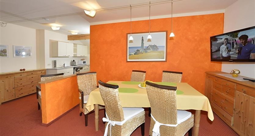Ein gemütlicher Essbereich mit einem großen Tisch und Stühlen aus Rattan. Die orangefarbene Wand und das dekorative Bild verleihen dem Raum eine einladende Atmosphäre.