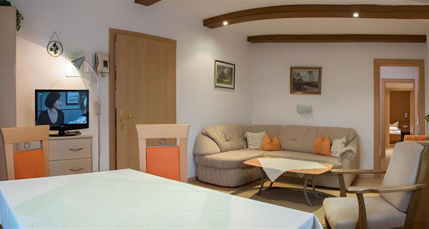 A cozy living room with a sofa and a table. There is a television and a dining area that is brightly lit.