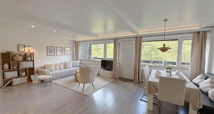 Een modern woonkamer met een gezellige zithoek en een groot raam. Het eetgedeelte heeft een elegante tafel en stoelen, omringd door natuurlijk licht.
