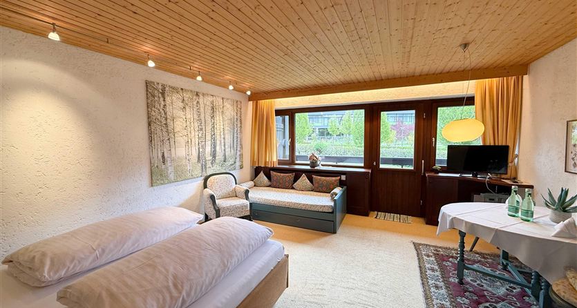 Een gezellige kamer met twee eenpersoonsbedden en een bank. Grote ramen bieden uitzicht naar buiten en zorgen voor veel licht.