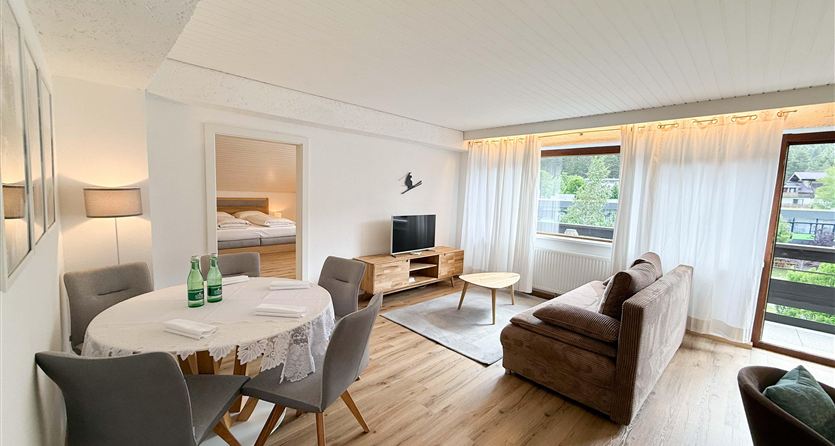 Een lichte woonkamer met een gezellige bank en een eettafel. Grote ramen zorgen voor veel daglicht en uitzicht op buiten.