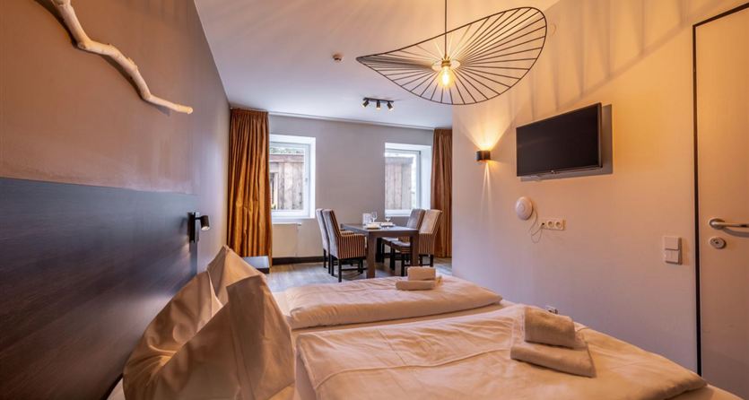A modern hotel room with two beds, a table, and chairs. Large windows provide plenty of natural light.