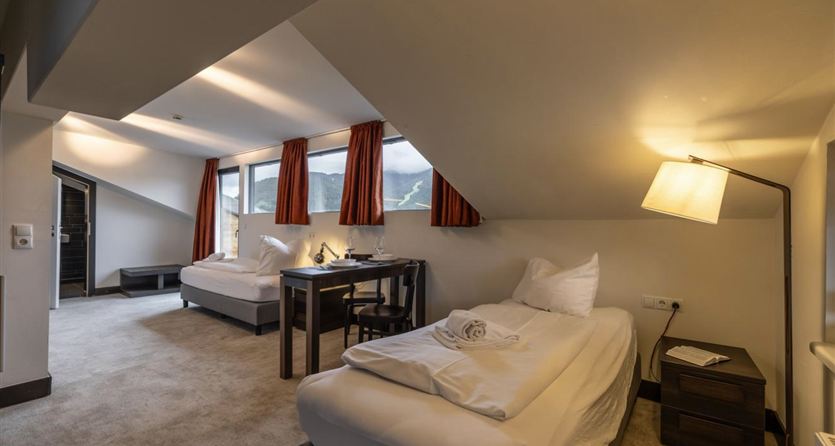 A cozy hotel room with two beds and a desk. Large windows offer a view of the surroundings.