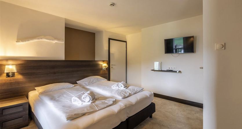 A modern hotel room with a double bed and two towels on the beds. There is a television and a decorative wall in neutral colors.