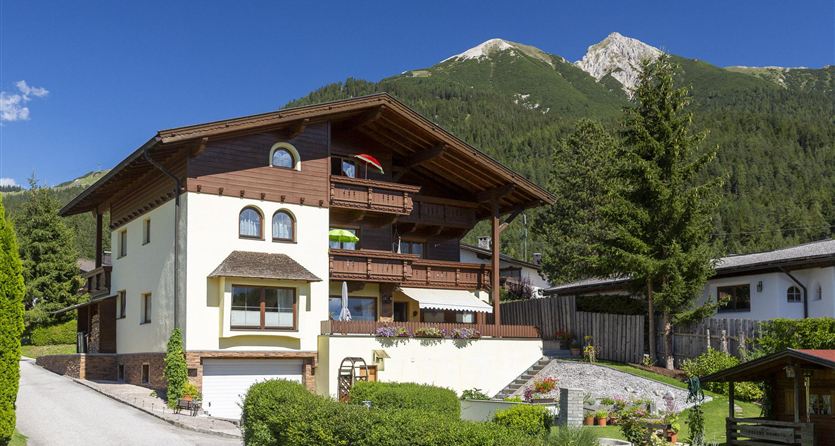 Een charmant huis in Alpenstijl omringd door groene bomen en bergen. De lucht is blauw en helder.