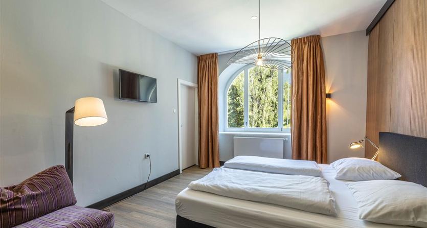 A modern bedroom with a double bed and a cozy sofa. Large windows let in plenty of light and offer a view of the greenery.